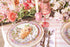 A styled table setting with floral ceramic plates, silver cutlery, a pink wine glass, and a heart-shaped pastry, surrounded by pink flowers and candles from oyster and arrow