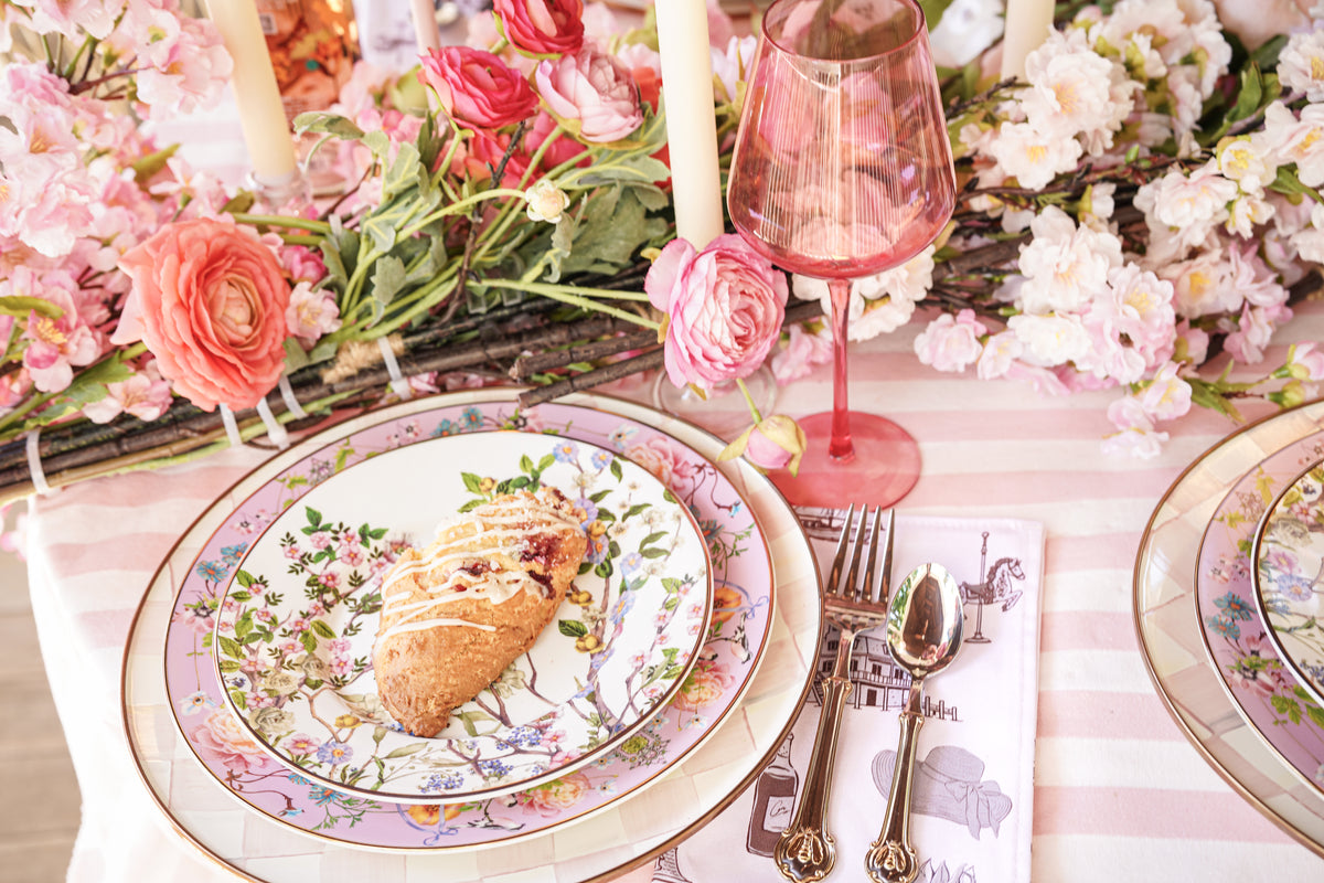 A styled table setting with floral ceramic plates, silver cutlery, a pink wine glass, and a heart-shaped pastry, surrounded by pink flowers and candles from oyster and arrow