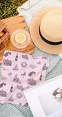 Pink patterned scarf with picnic items including a straw hat, basket, and glass on a blanket.