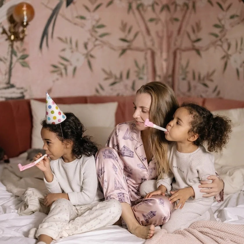 Two children on a bed blowing party horns and a mother with a decorative wall in the background