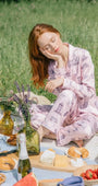 Woman in a floral dress sitting on a picnic blanket in a grassy field with food and drinks.