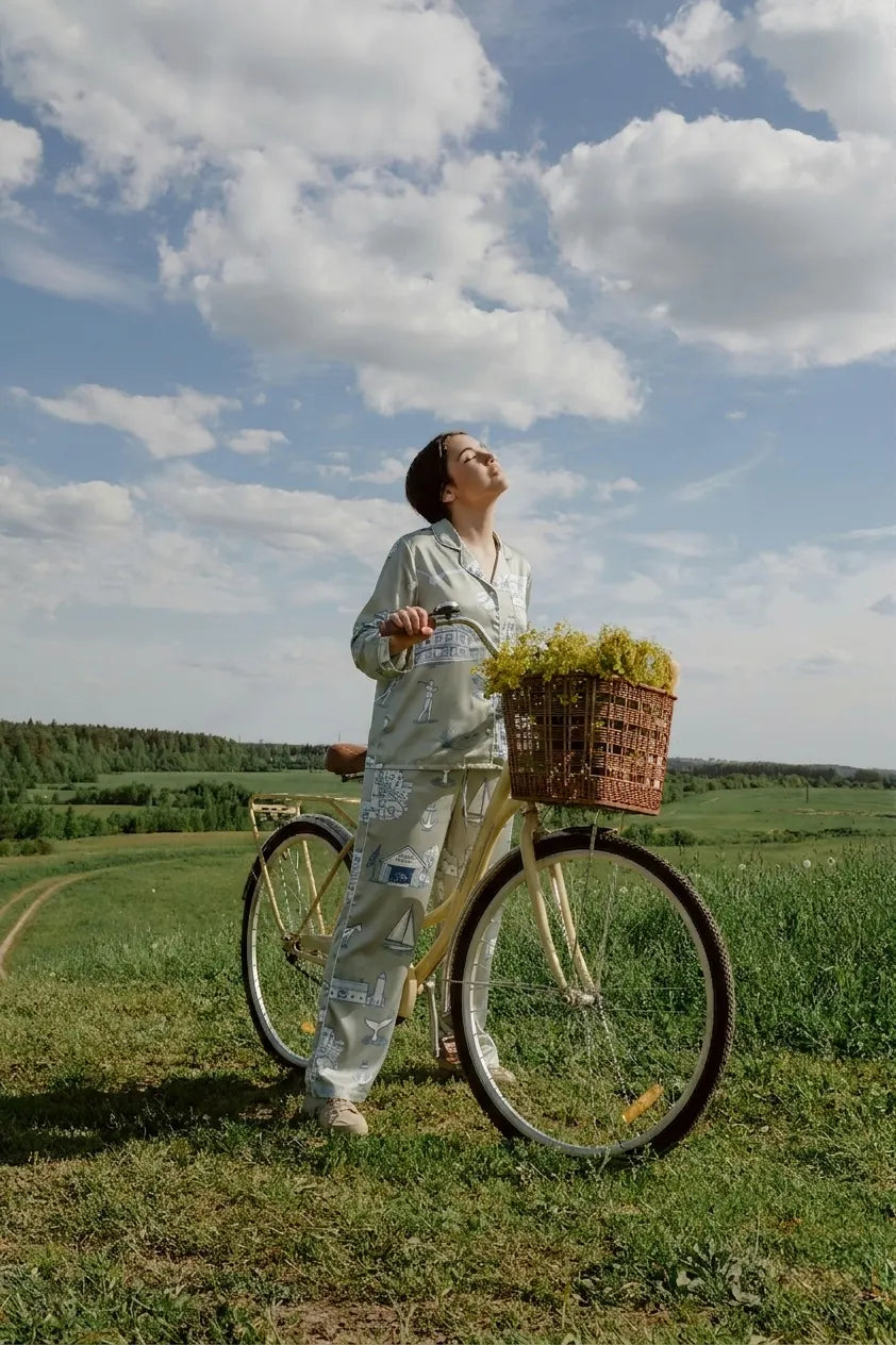 Woman in womens pajama set with button front standing by a bike in a field, enjoying the sunny sky.