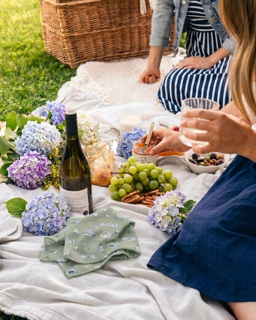 People enjoying a picnic with wine, food, and flowers on a blanket outdoors.