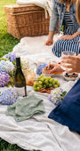 People enjoying a picnic with wine, food, and flowers on a blanket outdoors.