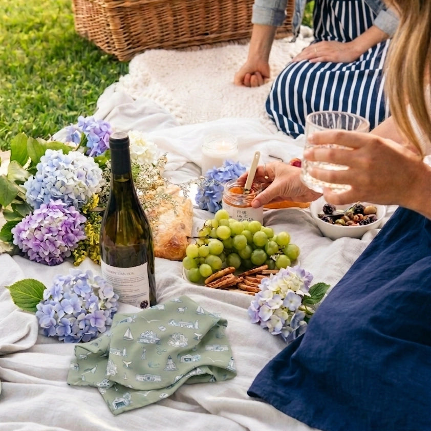People enjoying a picnic with wine, food, and flowers on a blanket outdoors.