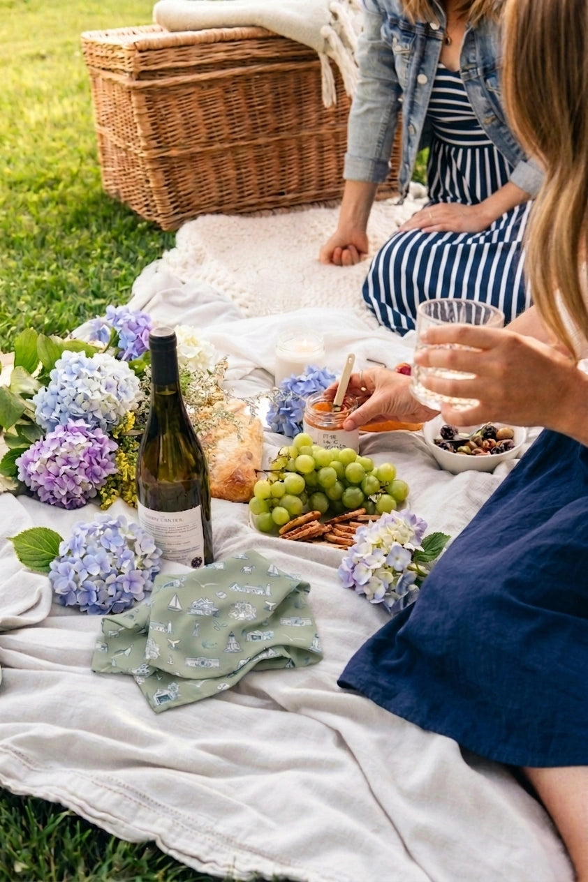 People enjoying a picnic with wine, food, and flowers on a blanket outdoors.