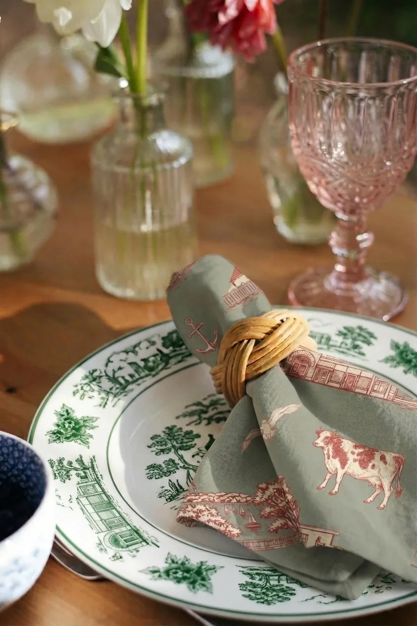 Decorative plate with a cow design, rolled napkin, and pink glass on a wooden table.