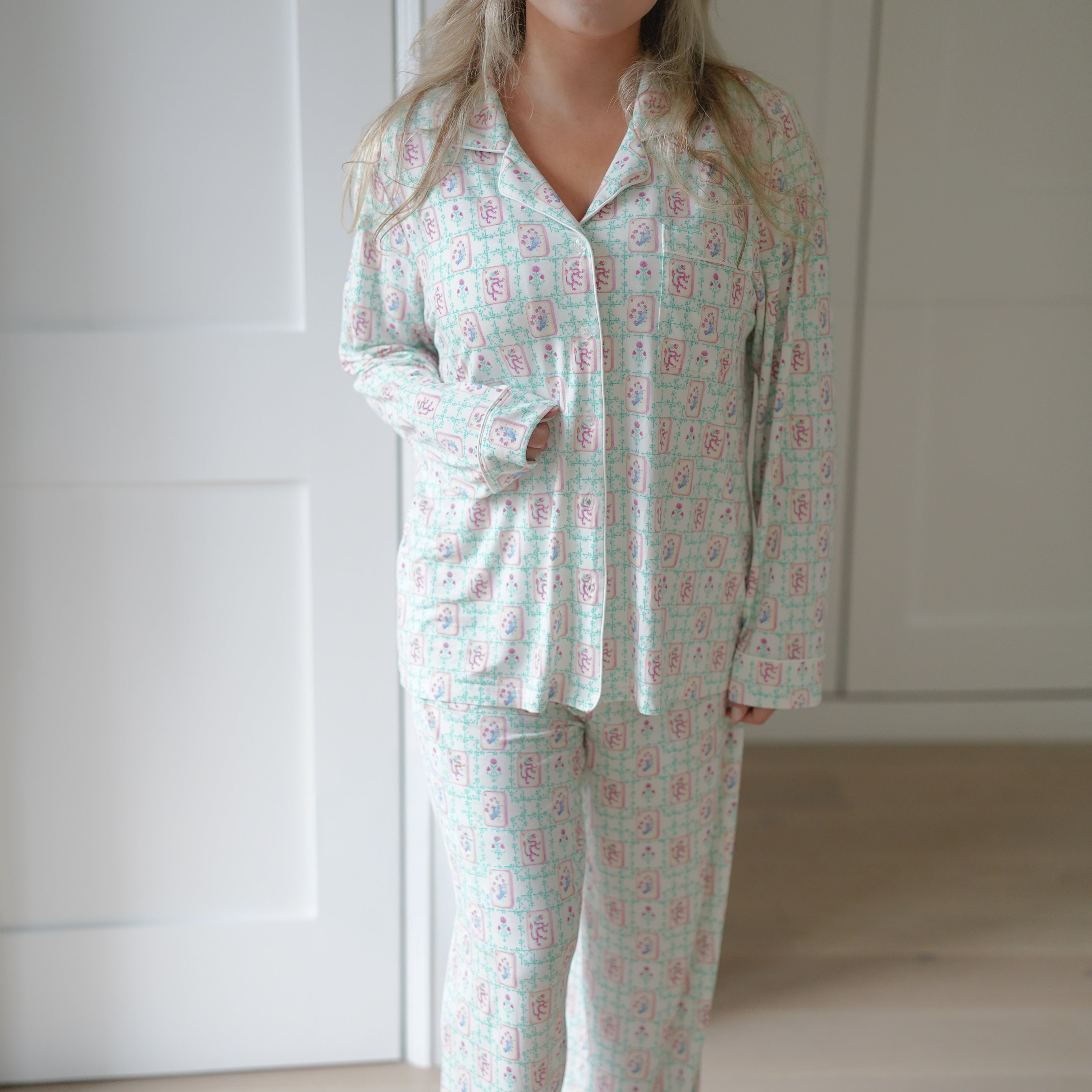 A woman wearing a matching Mahjong long-sleeve pajama set with a white base and a pink and teal floral pattern, standing in a brightly lit room with white paneled doors and light wood flooring.