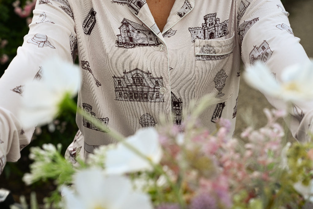 A close-up of a person wearing a cream-colored long-sleeve button-up toile pajamas with a black architectural house pattern from oyster and arrow, set against a soft-focus background of white and pink flowers.
