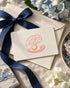 A cream-colored Oyster and Arrow gift card featuring the logomark styled on top of cream envelopes with a navy blue ribbon, surrounded by blue and white flowers on a linen surface.