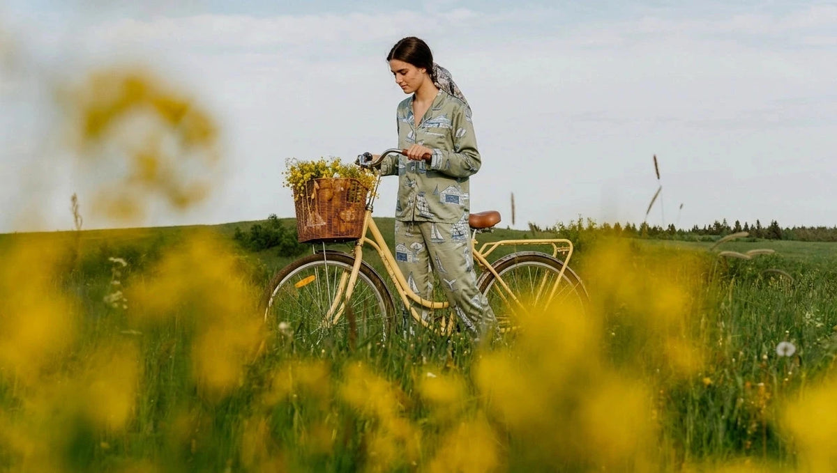 Woman with a bicycle in a field of yellow flowers wearing oyster and arrow coastal toile pajamas