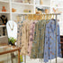 A clothing rack in a boutique displaying a row of long-sleeve button-up coastal toile bamaboo pajamas in soft pastel colors with hamptons area illustrations, set against a softly blurred shop interior with shelves and plants from Oyster and Arrow