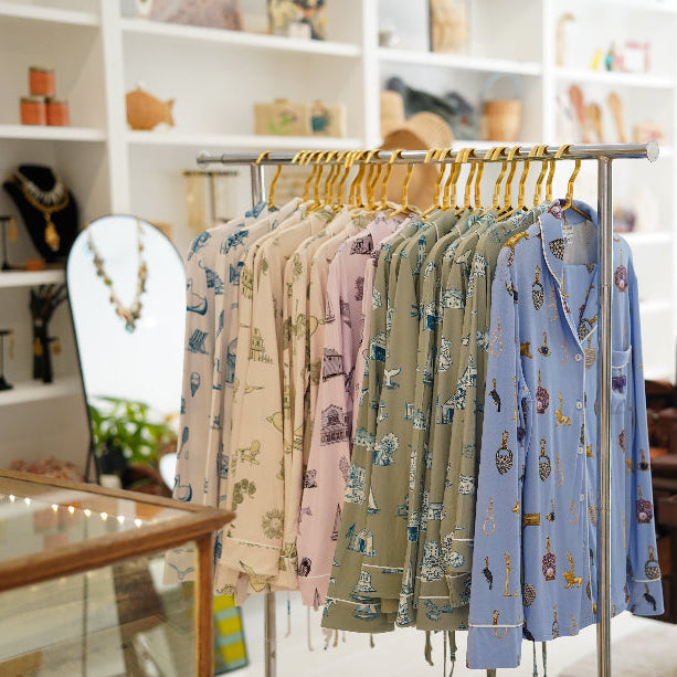 A clothing rack in a boutique displaying a row of long-sleeve button-up coastal toile bamaboo pajamas in soft pastel colors with hamptons area illustrations, set against a softly blurred shop interior with shelves and plants from Oyster and Arrow
