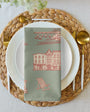 Dining table setting with a woven placemat, white plates, gold cutlery, and a decorative napkin.