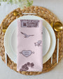 Table setting with a pink napkin featuring illustrations on a woven placemat.