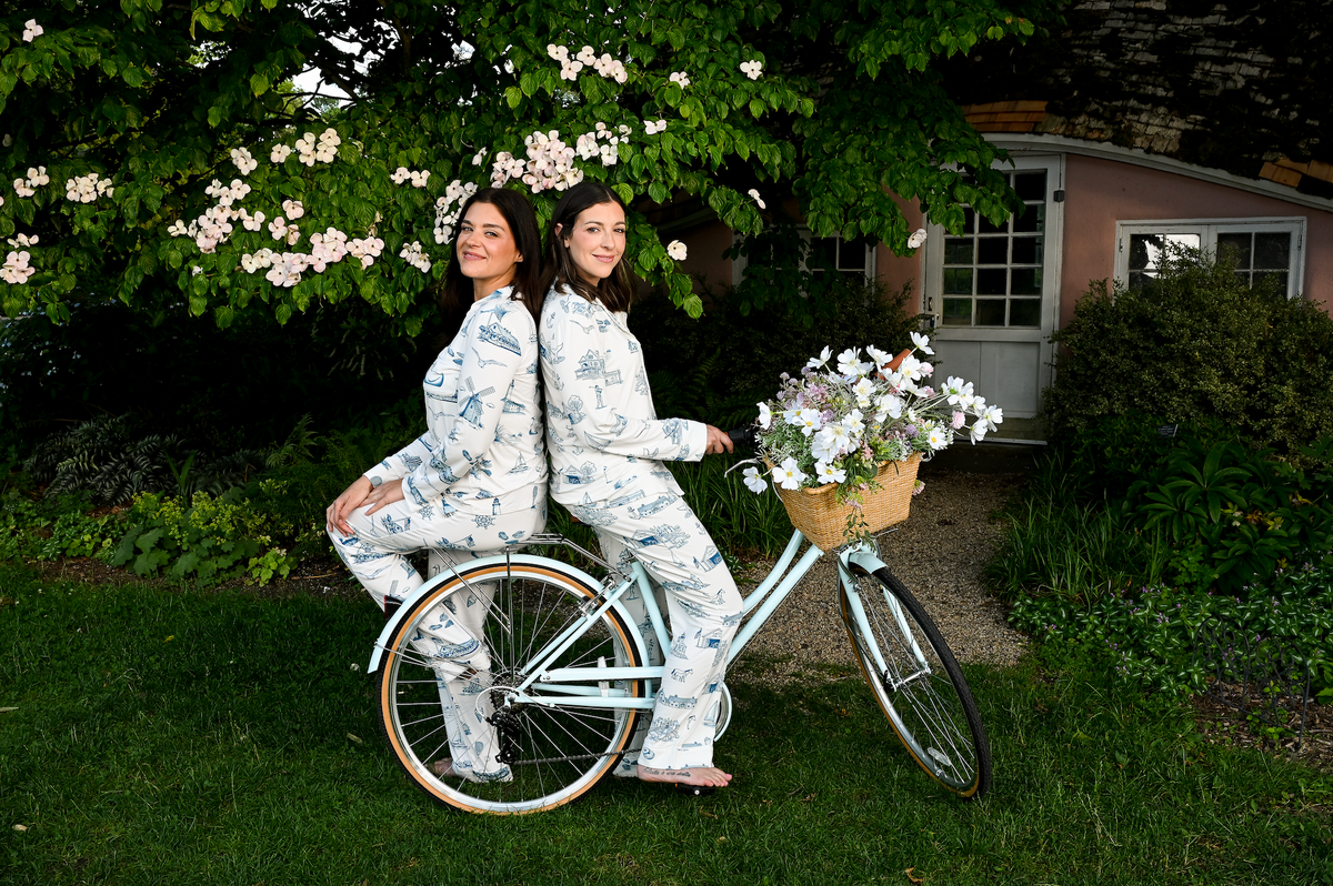 two girls sitting on a bicycle in the northeast with a pink house behind them wearing matching costal toile print bamboo pajamas from oyster and arrow